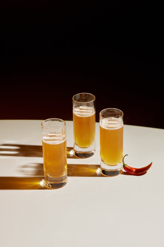 Three shot glasses, filled with orange-y, brown liquid. Contrast lighting. Small red chili pepper on the far right. 